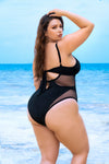 A woman wearing a one-piece swimsuit with transparent mesh cutouts, standing on the beach with the ocean in the background. The swimsuit has comfortable underwire support, with adjustable straps for a custom fit and a medium coverage bottom for comfort and style. The daring mesh cutouts provide a playful, flirty look. The inclusive sizing options ensure a perfect fit for all body types