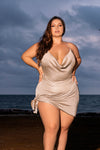 A woman wearing an asymmetrical swimwear dress with a draped cowl neck and side tie, standing on the beach with the ocean in the background. The shimmery and eye-catching design flatters all body types, and the dress comes in inclusive sizing. Get ready to turn heads and make a statement at the beach or pool with this one-of-a-kind look. 